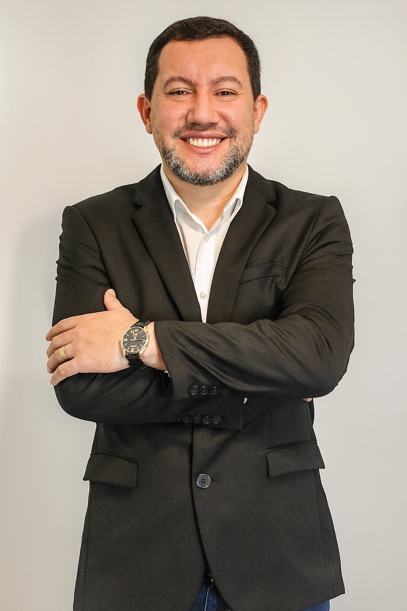 Edu Moraes sorrindo, de braços cruzados, com blazer preto e camisa branca